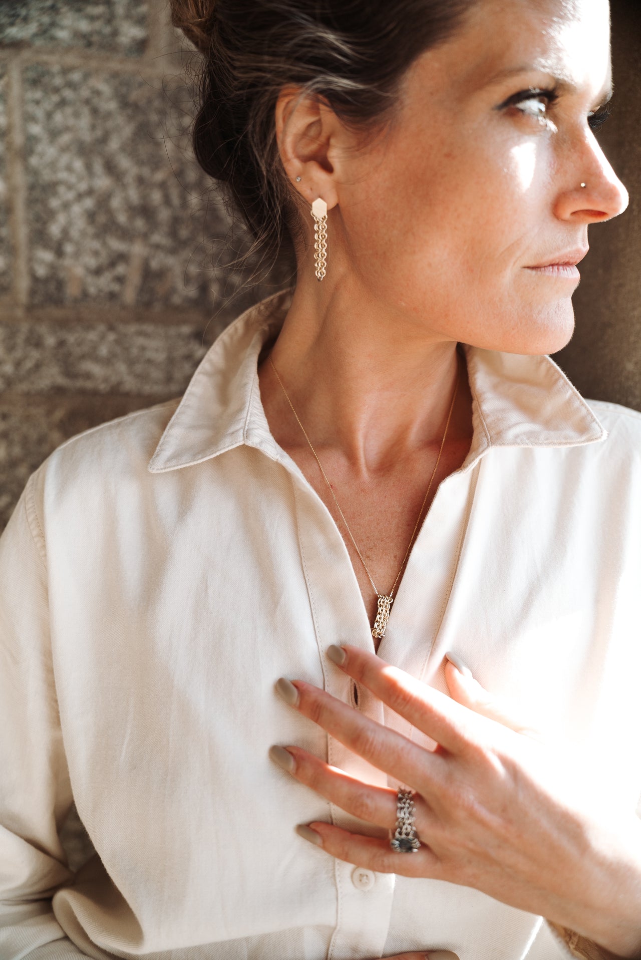 A person wearing a white shirt and displaying a long gold  chainmail earring with a hook back, part of the Moorland Glow collection.