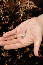 A close-up image of a hand holding a 'Stone Circle' white gold necklace with a pendant featuring a chainmail chain and a blue rectangular sapphire pendant, set in a white gold frame with yellow gold edging.