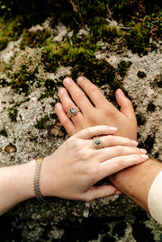 two modelled hands cross. A chainmail gold ring sits on the finger of both models. Gold chainmail and large sapphire stone rings. A gold chain link bracelet in yellow and white is positioned across one of the models wrists
